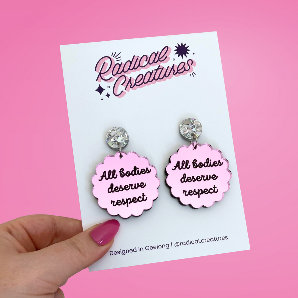Scalloped circle shaped dangle earrings with sparkly earring topper. Pink mirror with text "all bodies deserve respect"