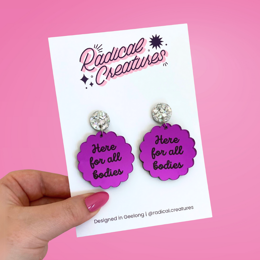 Scalloped circle shaped dangle earrings with sparkly earring topper. Purple mirror with text "here for all bodies"