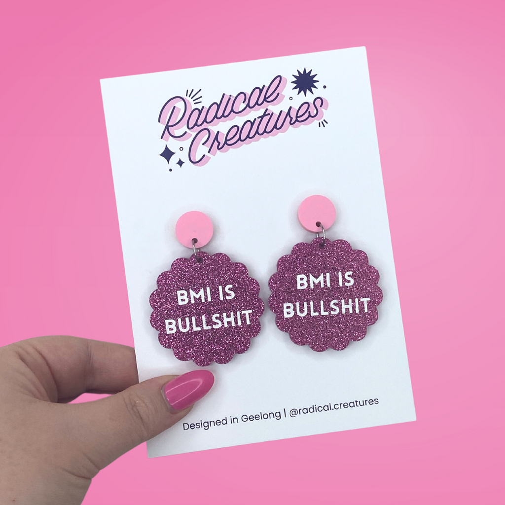 Pink glittery acrylic earrings in scalloped circle shape with white block letters that say "BMI is bullshit"