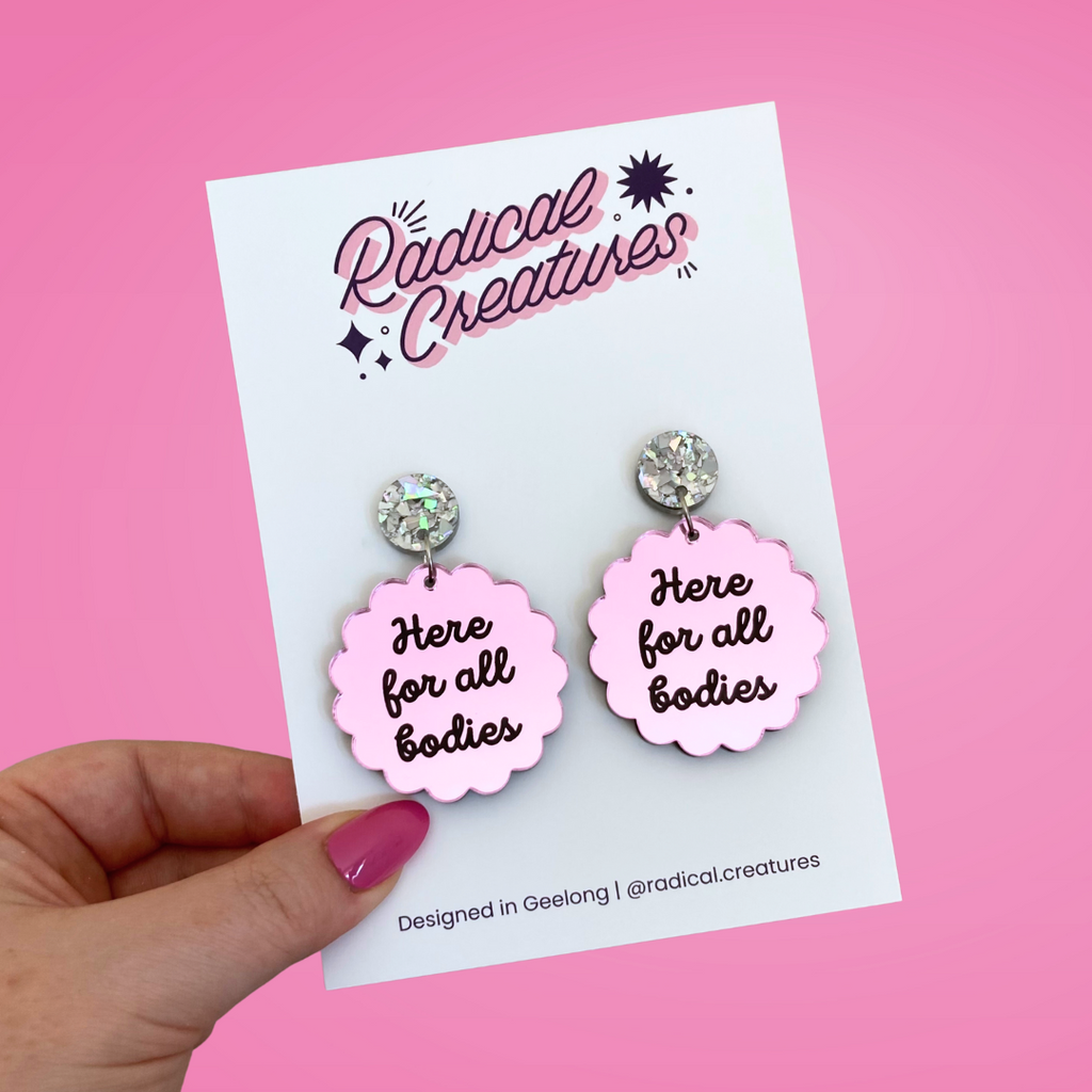 Scalloped circle shaped dangle earrings with sparkly earring topper. Pink mirror with text "here for all bodies"