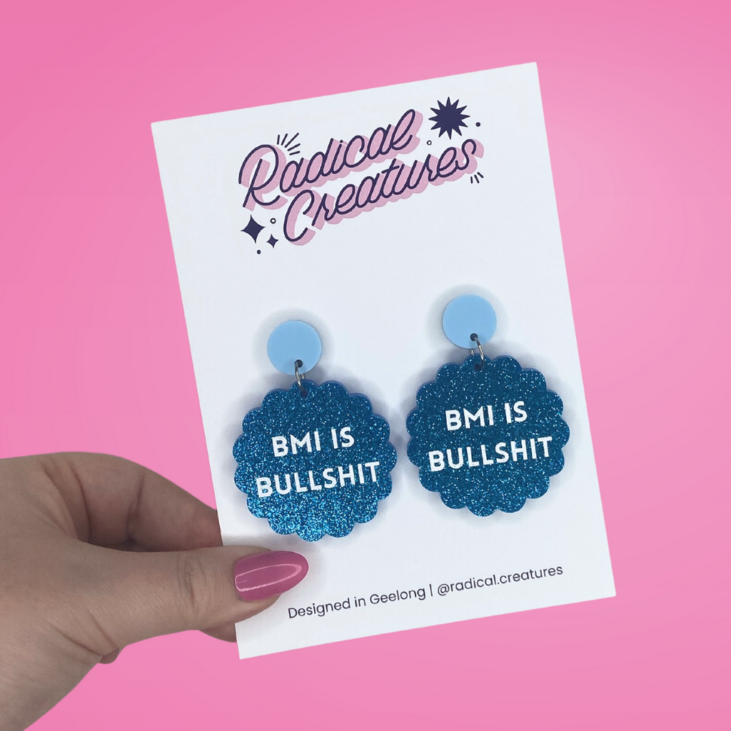 Glittery blue acrylic earrings in scalloped circle shape with white block lettering saying "BMI is bullshit"